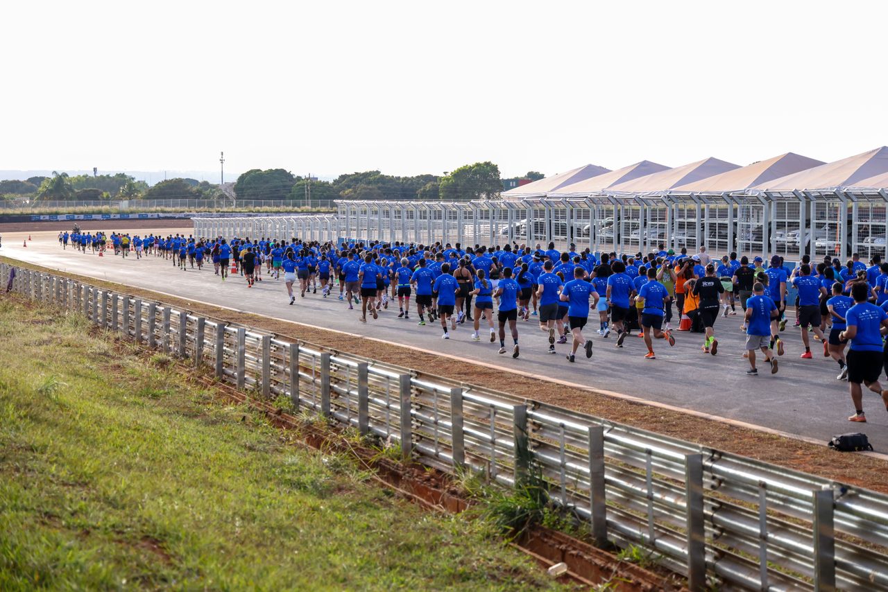 Um grande grupo de corredores vestindo camisetas azuis participa de uma prova de atletismo em uma pista ao ar livre.
