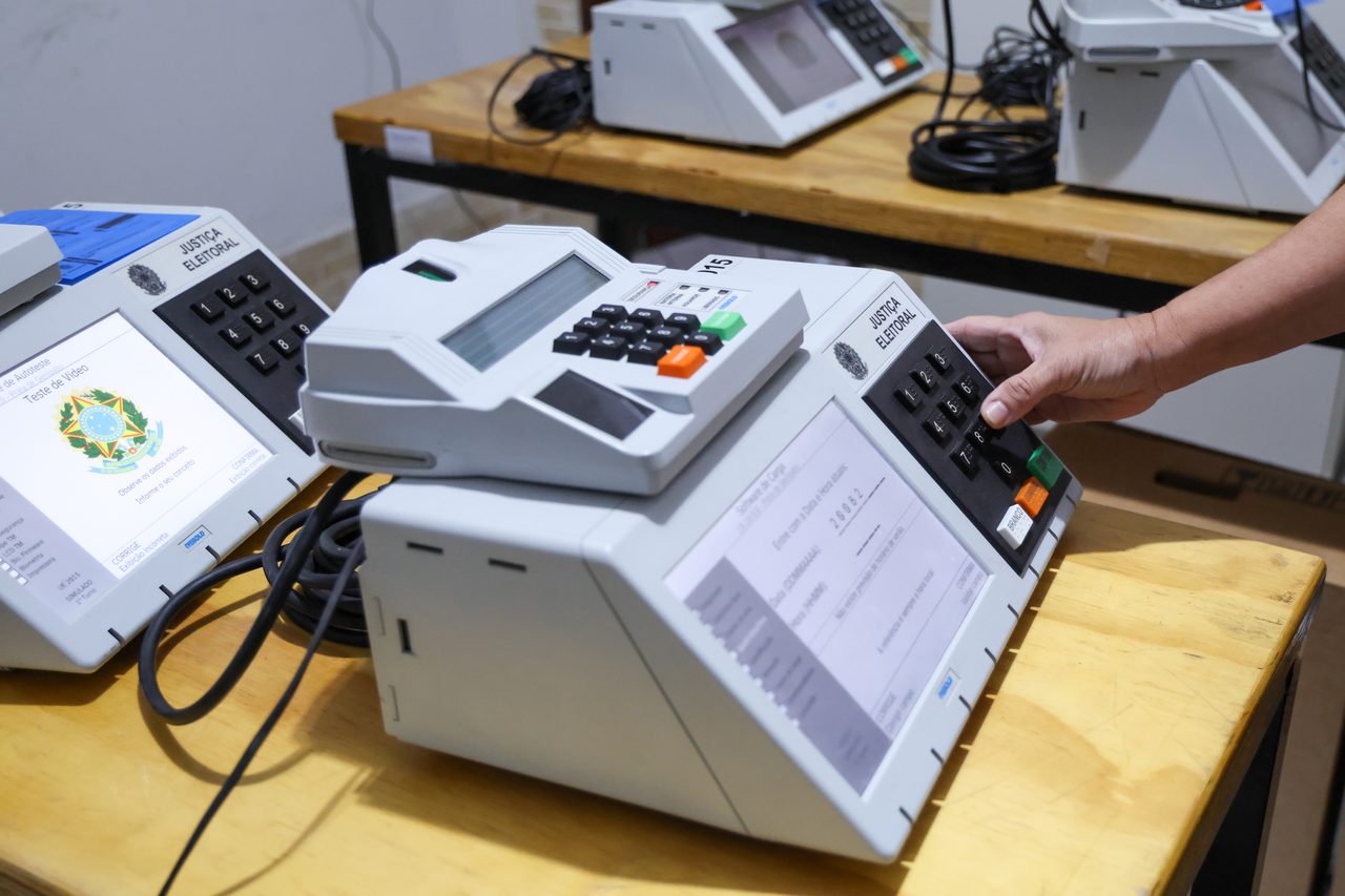 Close-up de uma mão operando uma urna eletrônica da Justiça Eleitoral brasileira sobre uma mesa de madeira.