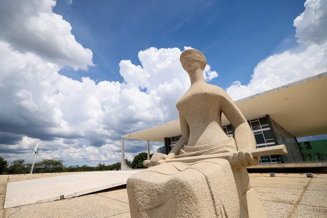 Uma escultura de pedra em formato humano sentada em uma praça, com um edifício de arquitetura moderna e céu nublado ao fundo.