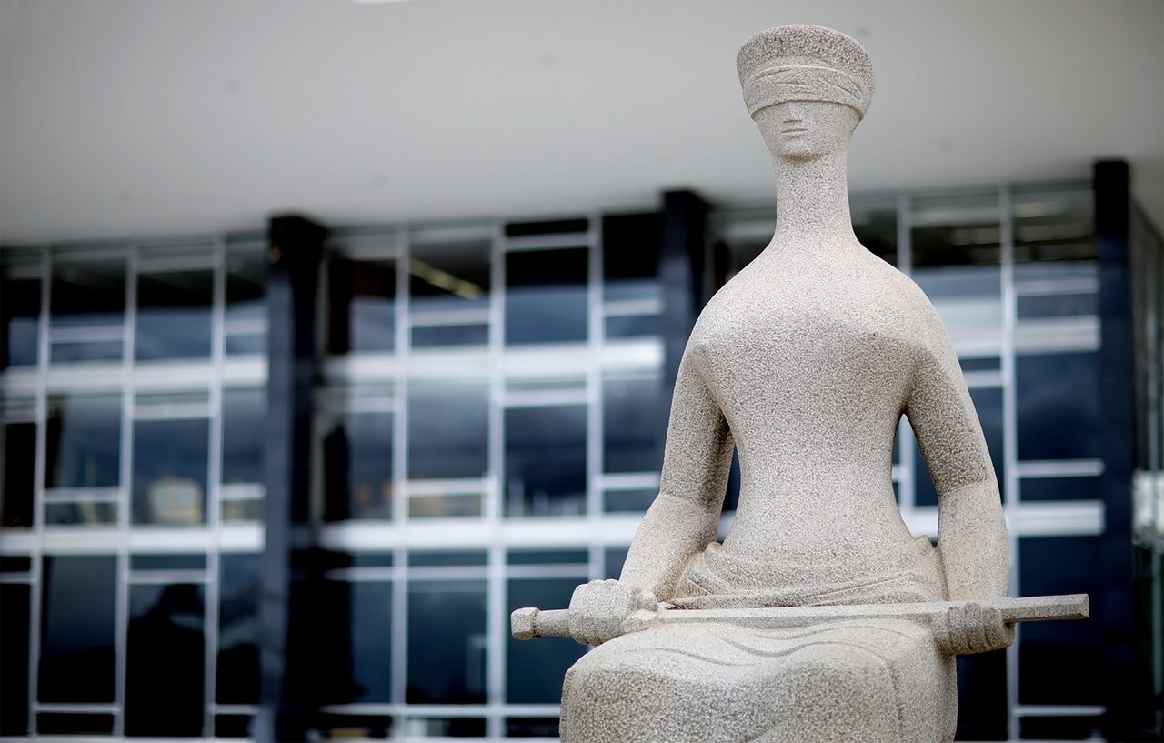 Estátua de pedra representando a justiça com os olhos vendados e segurando um objeto, posicionada em frente a um edifício moderno com janelas de vidro.