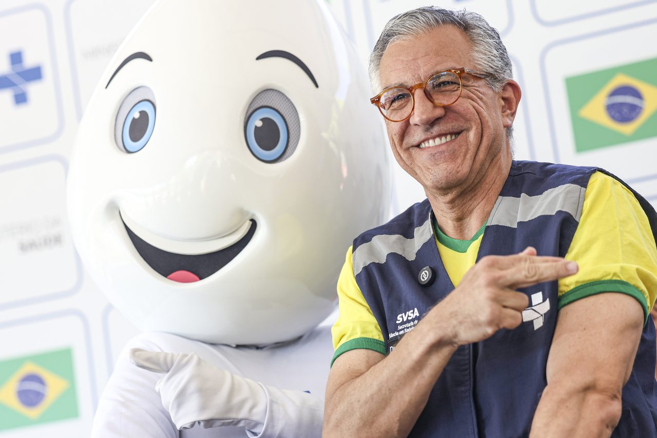 Homem sorridente ao lado de um mascote de campanha de vacinação da Secretaria de Vigilância em Saúde. Na foto: Alexandre Padilha.